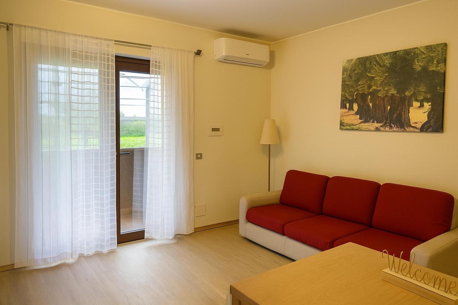 The living room: red sofa, window with light curtains and a painting of olive trees