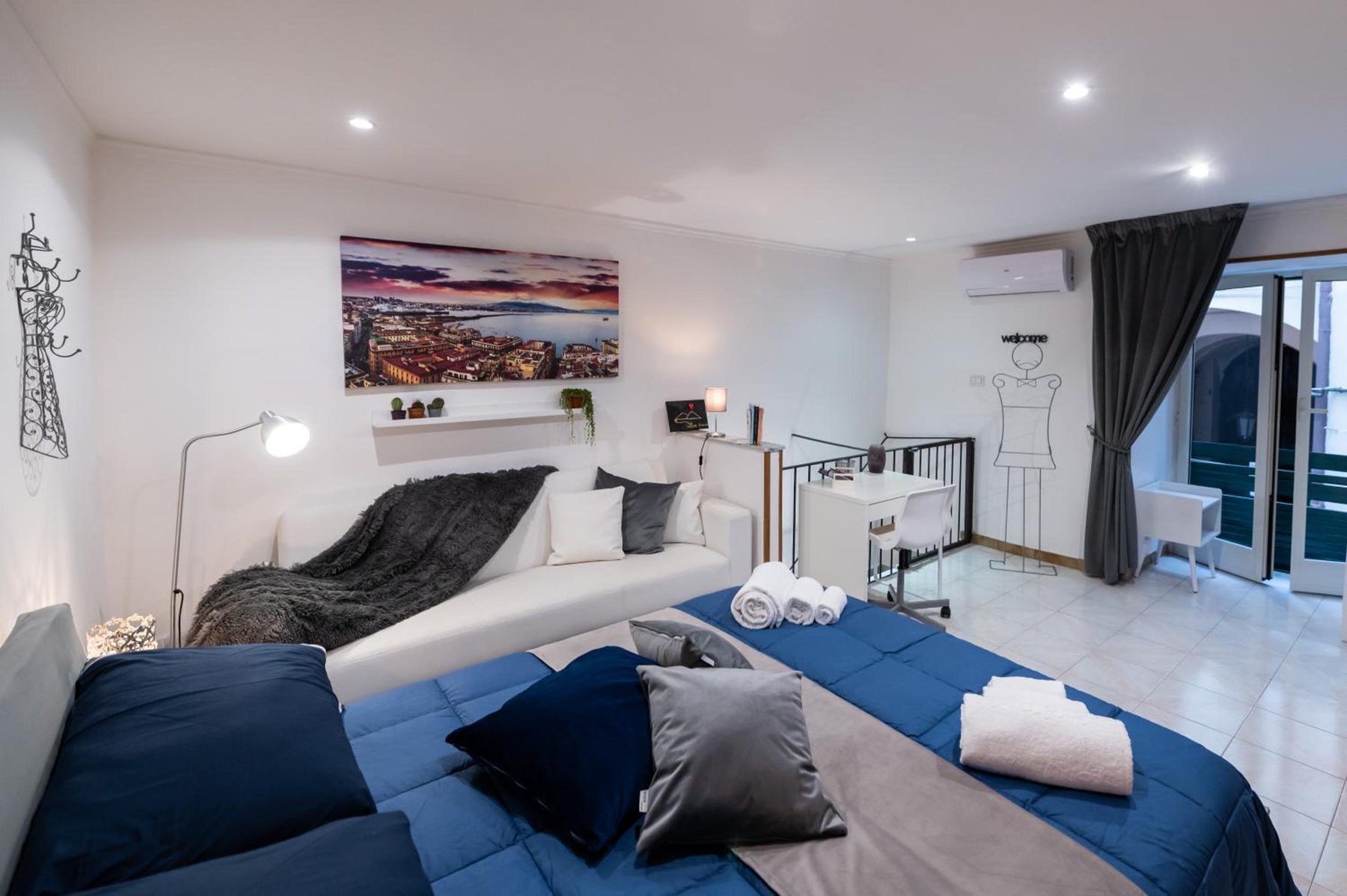 Room with a blue bedspread, grey cushions and a print of the Bay of Naples above the bed