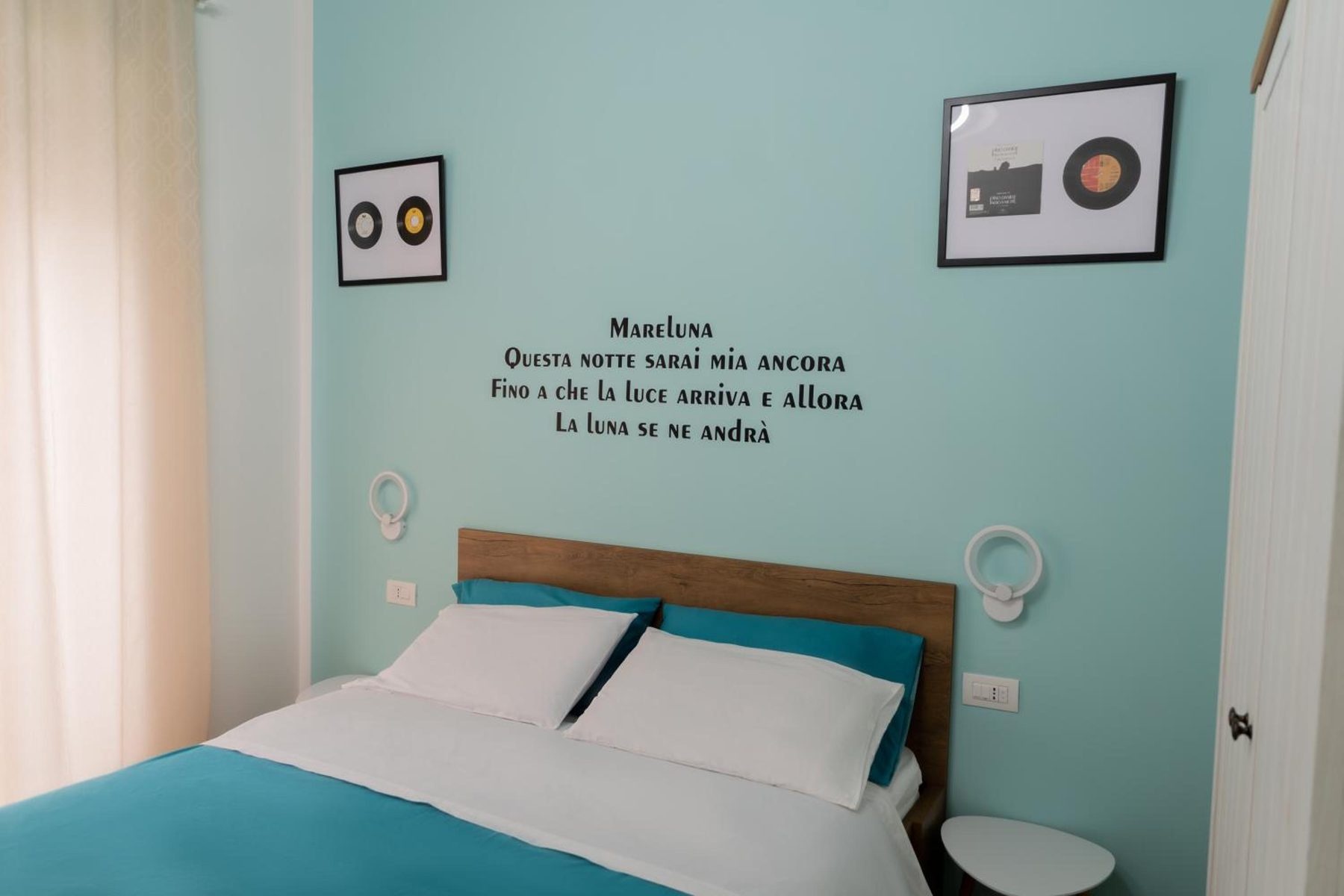 Mareluna: bed with a teal bedspread, two framed records and the lettered wall